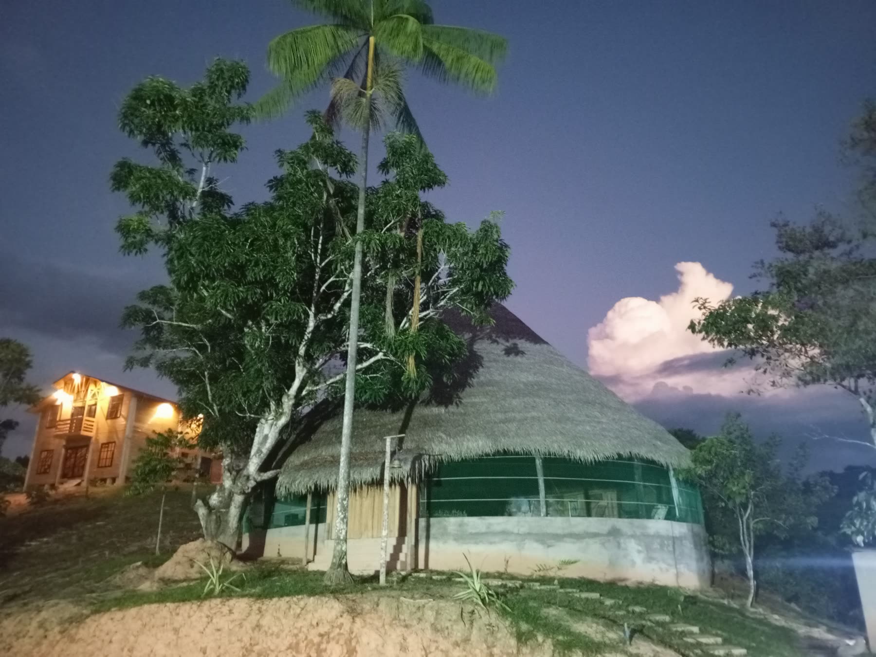 Maloca de techo de palma al anochecer, con un bungalow iluminado a lo lejos